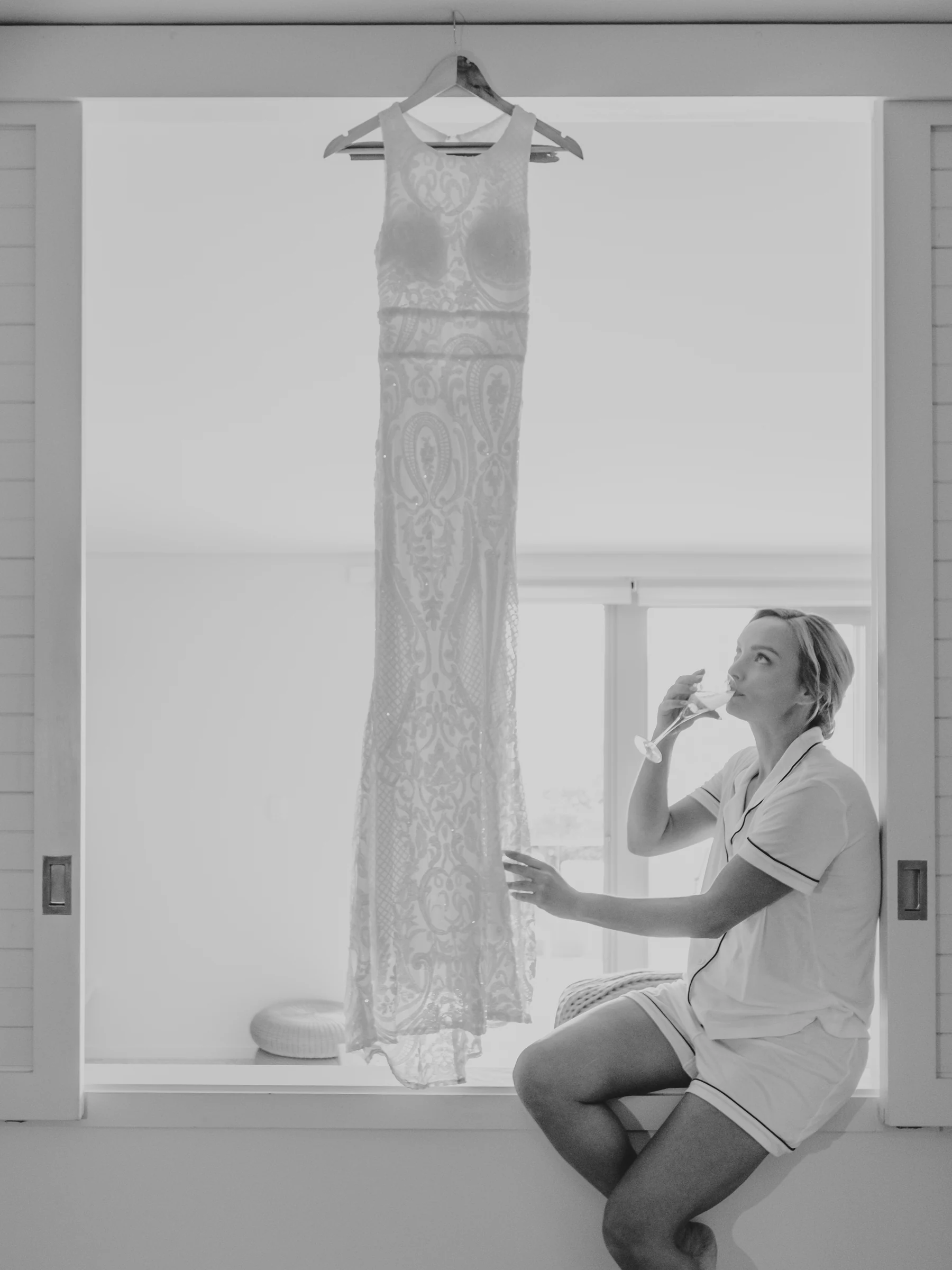 Black and white portrait of Victoria sipping champagne with her wedding dress hanging in the window behind her