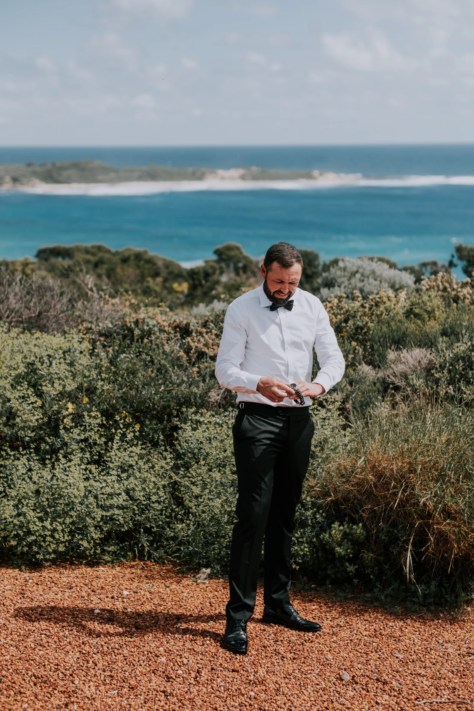 Luck adjusting his cufflinks in his white shirt and bow tie on the gravel path, ocean and headland behind him at Injidup Beach
