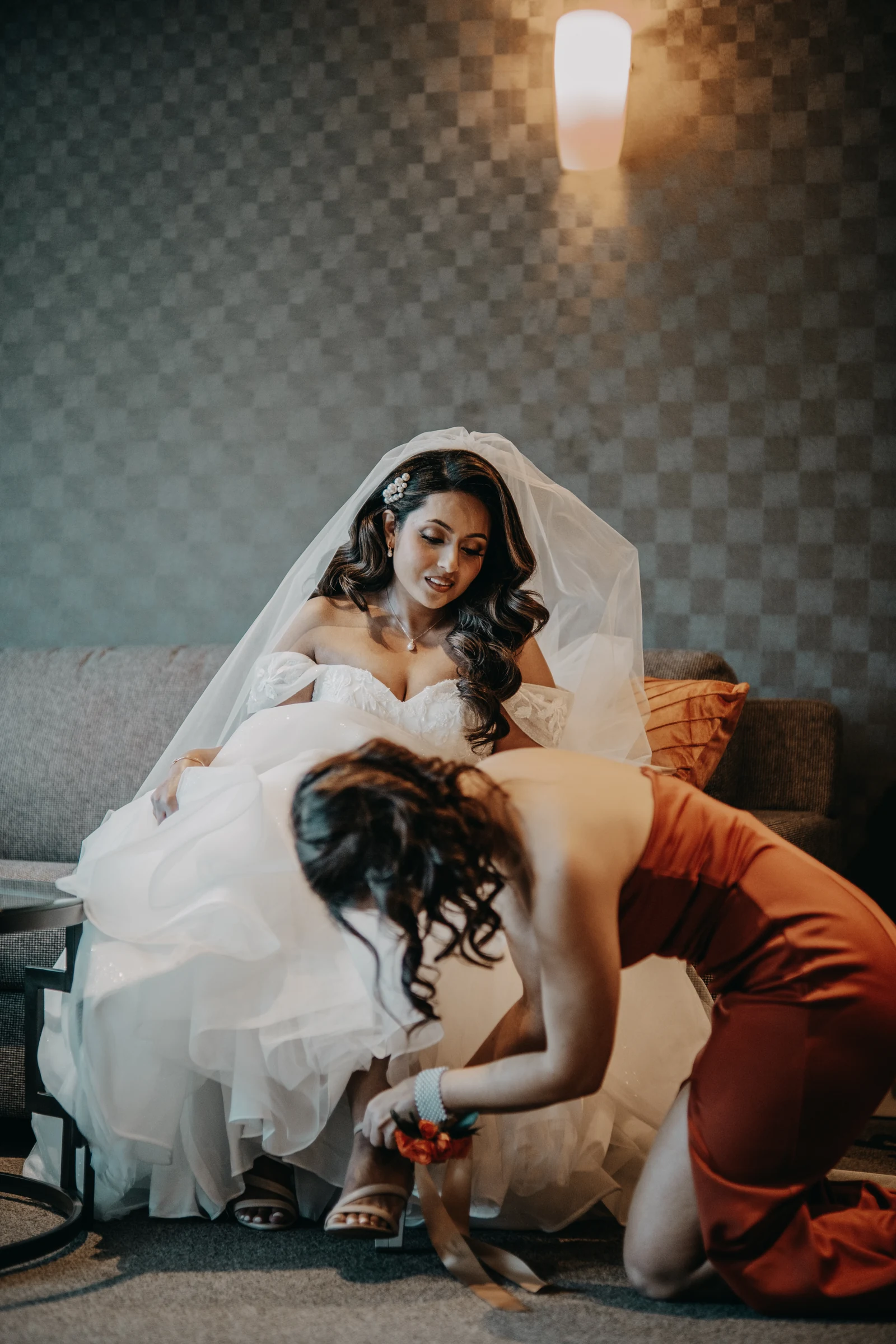 Betsy relaxing on a couch in her robe as her maid of honour adjusts her wedding shoes