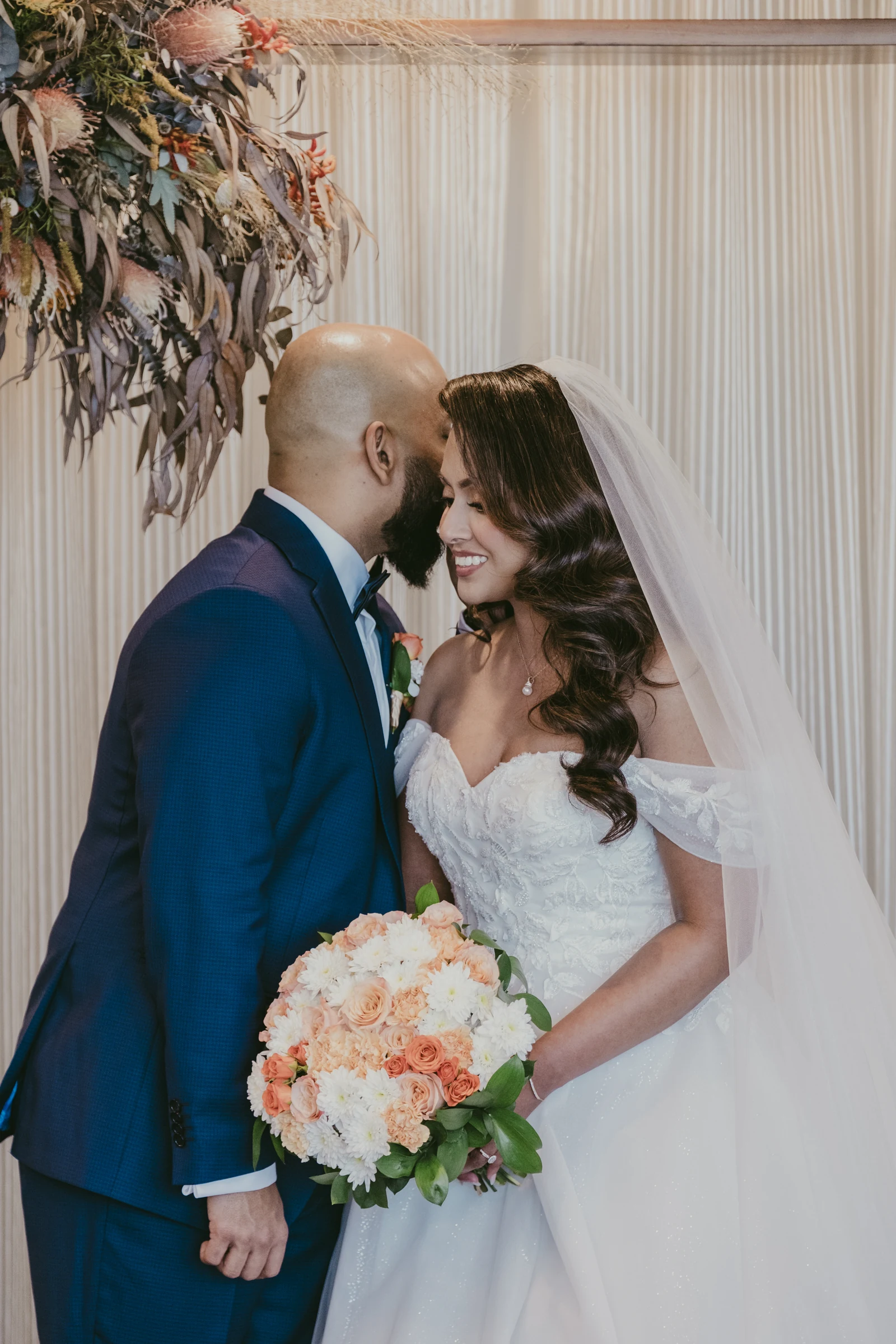 Betsy and Jibin embracing beneath a floral arch just after being pronounced married