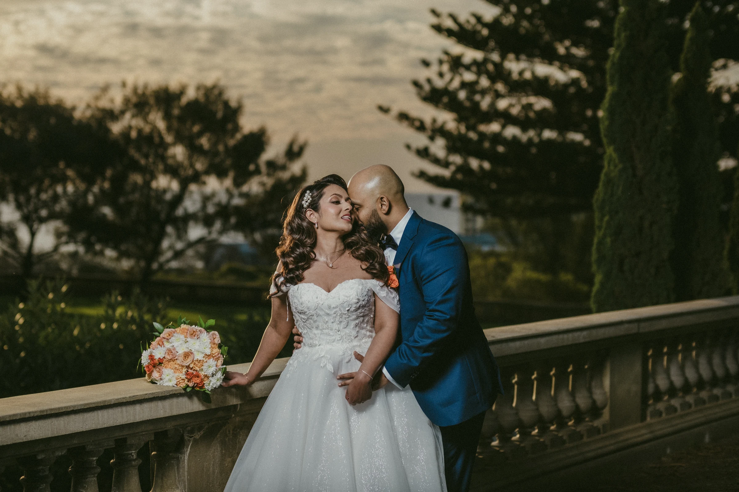 Jibin kissing Betsy's cheek as she holds her bouquet on the heritage balustrade at Cottesloe Civic Centre under a glowing dusk sky