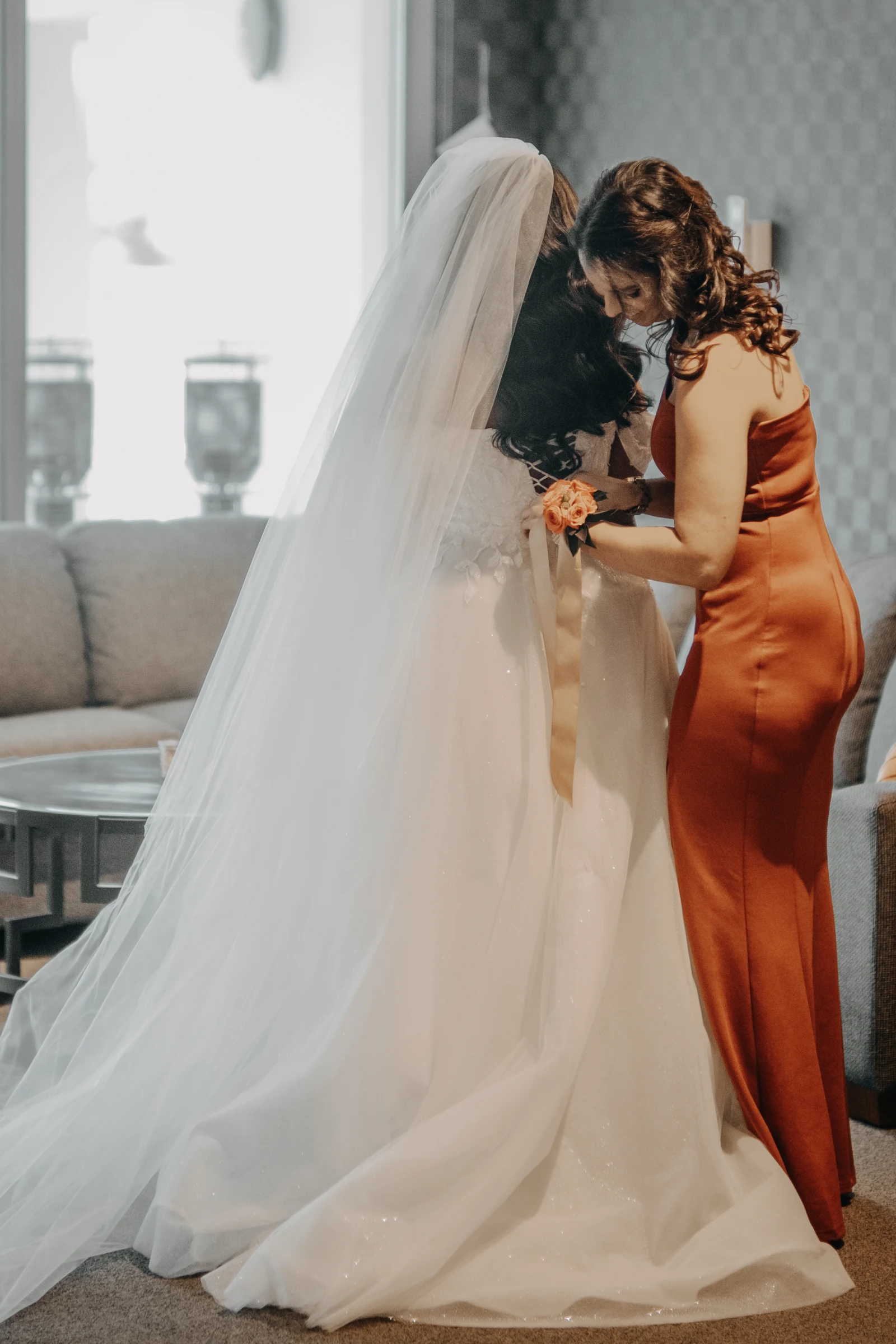 Betsy's maid of honour helping zip up her wedding dress by the window