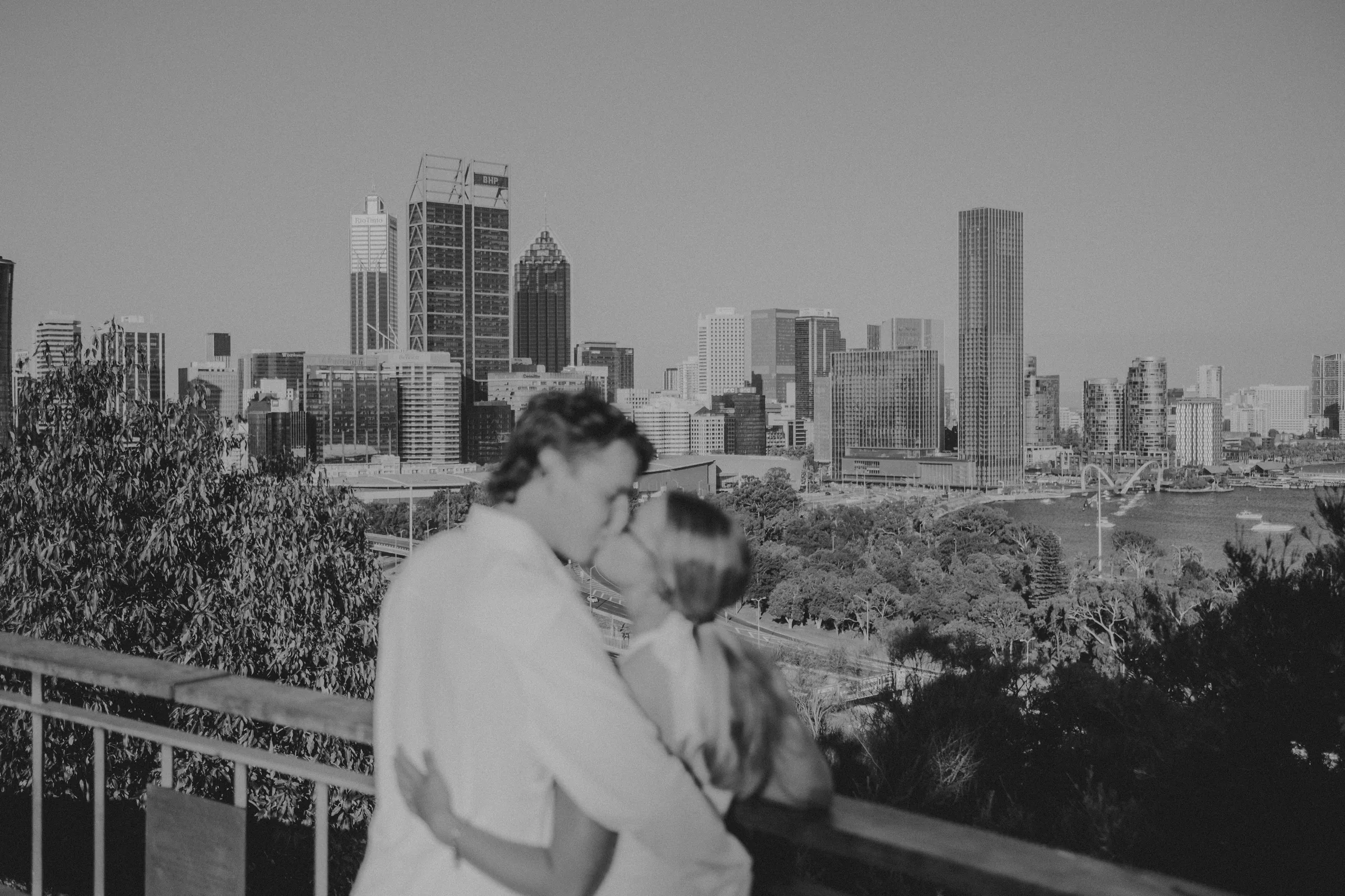 Black and white photo of Kayla and Brett embracing at a Kings Park lookout with the Perth city skyline in the background