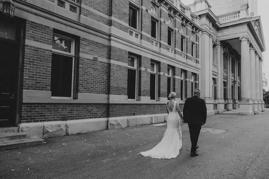 Black and white photo of a bride and groom walking beside the heritage Supreme Court Gardens building, Perth CBD wedding photography