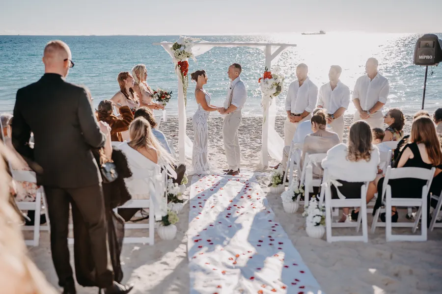 Wide shot of Katia and Sean holding hands at a white timber arbour on the sand, guests seated either side, Coast Port Beach