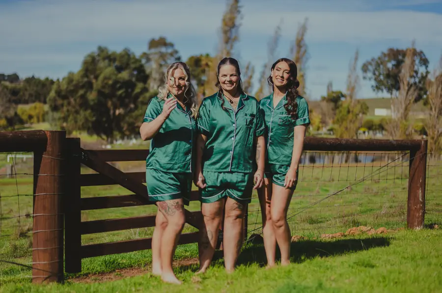 Bridesmaids in green satin pyjamas laughing together at a farm gate, Dalmore Farm wedding morning