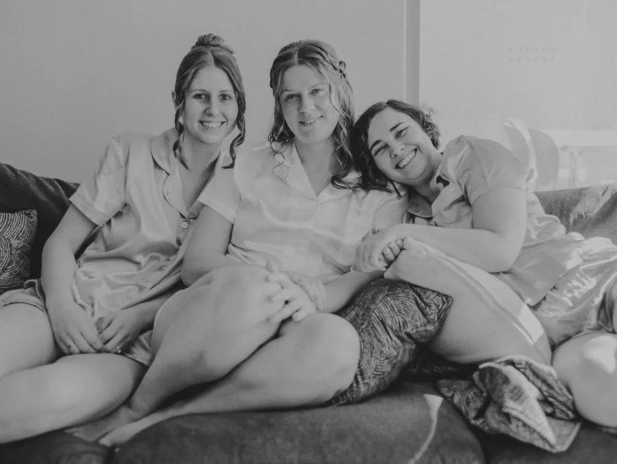 Three bridesmaids in satin pyjamas laughing together on the couch getting ready for the wedding at The Barn, Hopeland