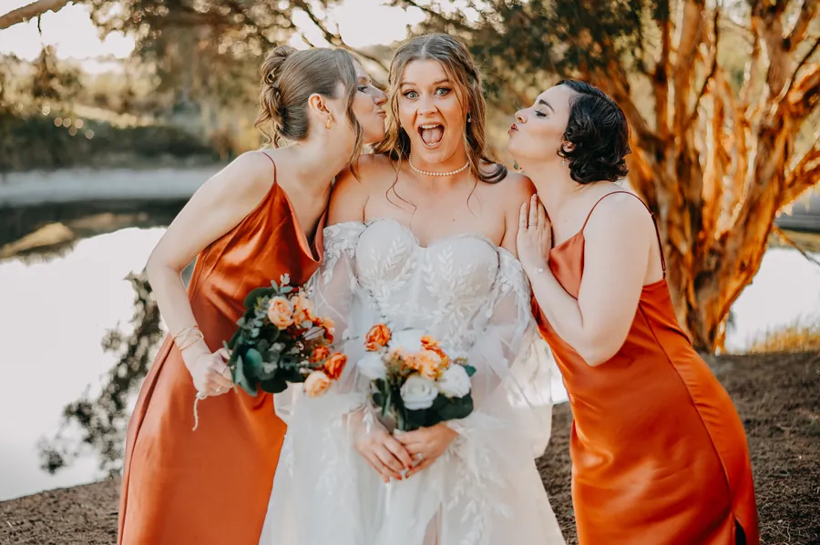 Bride laughing in shock as two bridesmaids in burnt-orange dresses kiss her cheeks by the lake
