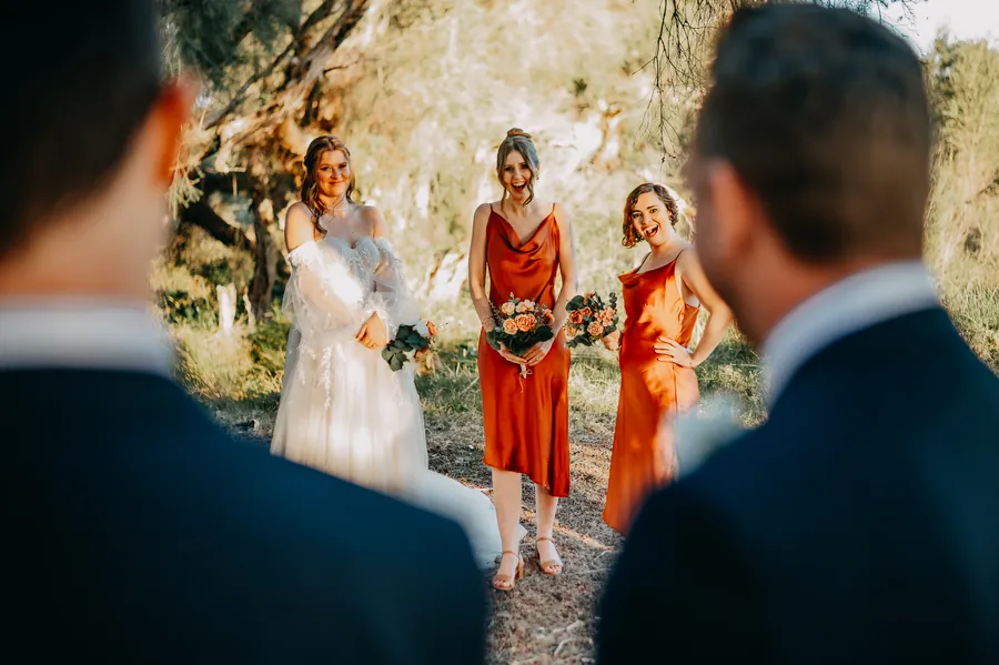 Bride and bridesmaids laughing candidly together, framed between two groomsmen in the foreground