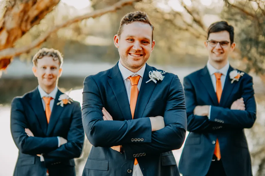 Groomsmen in navy suits and orange ties standing arms crossed and smiling at golden hour
