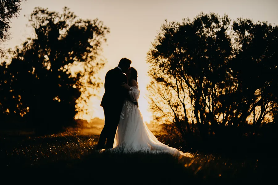 Silhouette of the bride and groom kissing beneath the trees as the sun sets over Hopeland