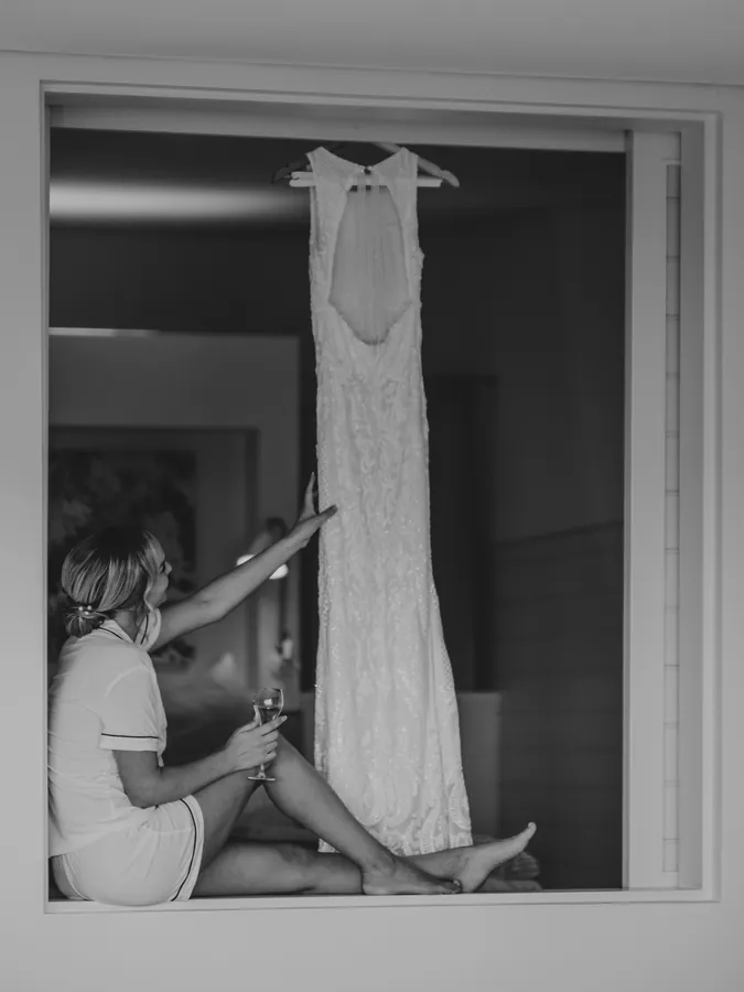 Black and white portrait of Victoria reaching for her wedding dress hanging in the window