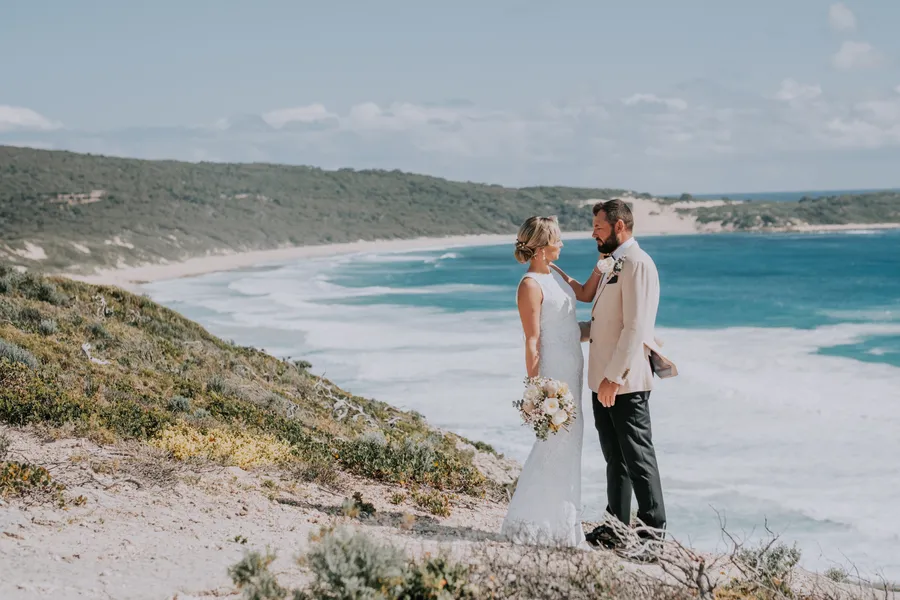 Victoria and Luck face to face on the dune top before the ceremony, Victoria holding her bouquet, Injidup Beach coastline behind them