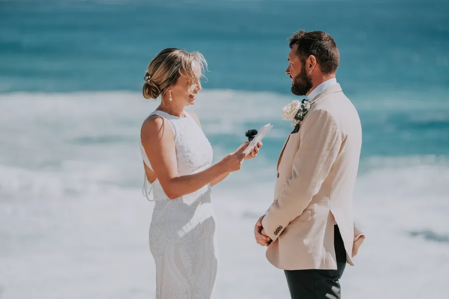 Victoria laughing as she reads her vows to Luck, ocean waves behind them