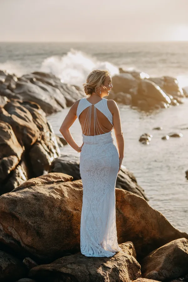 Victoria looking back over her shoulder in her open-back dress as a wave crashes on the rocks behind her, golden hour