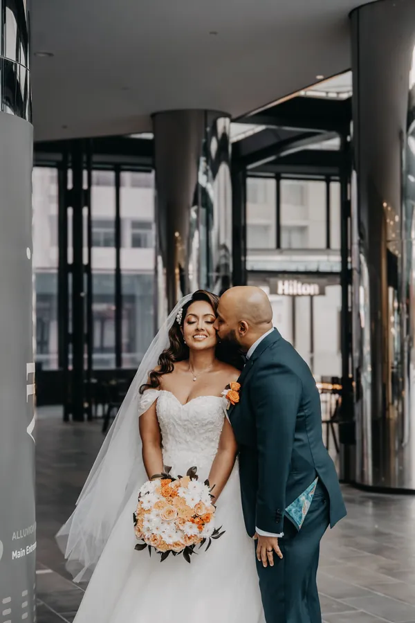 Jibin kissing Betsy on the cheek among the steel columns of a modern CBD walkway